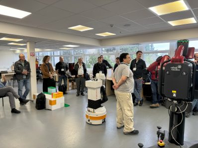 A group of people standing and listening to one person standing in the center with a robot standing next to them.