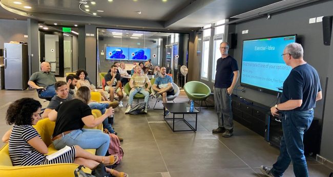A room in which a group of people sit on chairs while two people stand near a screen and talk.